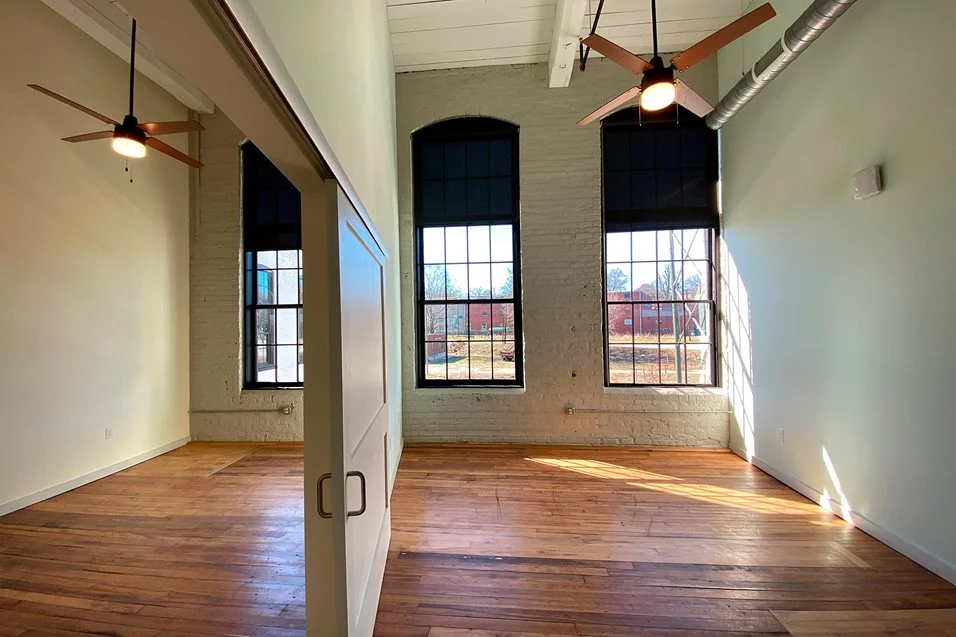 View inside of apartment with wood floor, white plaster and brick walls, large windows with light streaming in
